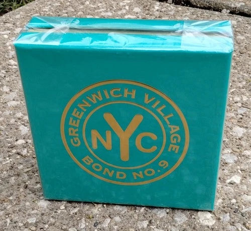 A square turquoise box wrapped in clear plastic, featuring a gold circular logo that reads 'GREENWICH VILLAGE NYC BOND NO. 9', placed on a rough concrete surface.